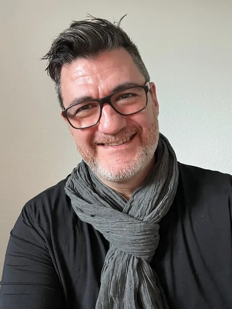 Portrait of Tony Wood, smiling, wearing glasses and a grey scarf.